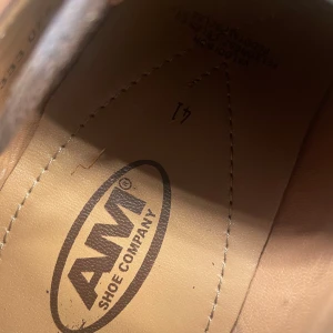 Bruna skinnskor från AM Shoe Company - Snygga bruna skinnskor från AM Shoe Company med klassisk design. Skorna har snörning, perforerade detaljer på sidorna och tåpartiet samt kontrastsömmar. Perfekta för dig som vill ha en stilren look med en modern touch.