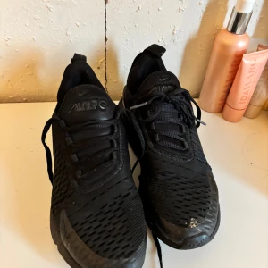 Svarta Nike Air Max 270 sneakers - Säljer ett par helsvarta Nike Air Max 270 sneakers med mesh-överdel och synlig Air-enhet i hälen. Skorna har snörning och en sportig vibe, perfekt för dig som gillar streetstyle. Snygg och clean design med diskreta Nike-loggor på tungan och sidan.