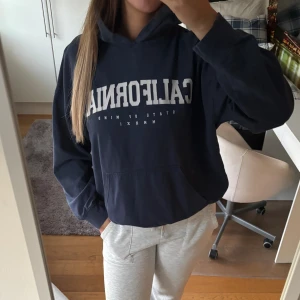 Mörkblå hoodie med tryck CALIFORNIA - Säljer en mörkblå hoodie med vit text 'CALIFORNIA' på bröstet. Tröjan har huva, känguruficka och en relaxed, lite oversized passform. Perfekt för chill dagar och enkel att styla med jeans eller mjukisbyxor.