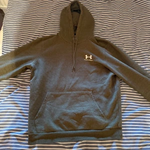 Grön hoodie från Under Armour - Säljer en grön hoodie från Under Armour i storlek M. Tröjan har huva med snörning, stor magficka och en vit logga på bröstet. Perfekt för dig som gillar en enkel och sportig stil. Materialet är mjukt och skönt, troligtvis bomull och polyester.