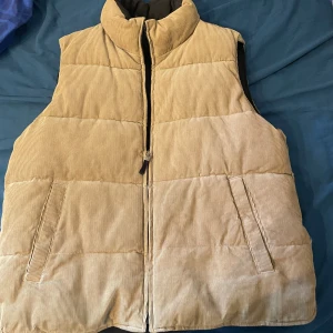 Beige quiltad dunväst i manchester - Säljer en beige dunväst med quiltad design och mjukt manchestertyg. Västen har hög krage, två snedställda fickor fram och dragkedja hela vägen. Insidan är svart och har en extra ficka med dragkedja. Perfekt för lager-på-lager under kyliga dagar.