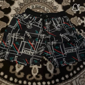 Svarta Lacoste shorts med mönster - Snygga svarta shorts från Lacoste med grafiskt mönster, logga och krokodiler i vitt, rött och grönt. Elastisk midja med snörning för skön passform. Perfekta för sommaren och chill dagar.