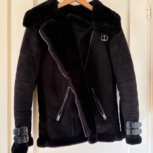 Svart jacka med spännen - Snygg svart fejk pälsjacka med stora slag, dragkedjor och coola spänndetaljer vid handlederna. Jackan har mjukt foder och två snedställda fickor med dragkedja. Perfekt för dig som vill ha en varm och trendig look under kalla dagar. Jackan är i bra skick förutom att den är sliten på insidan av fickorna.