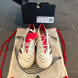 Adidas Predator Elite FG fotbollsskor - Säljer ett par Adidas Predator Elite FG fotbollsskor i vitt med svarta och röda detaljer som endast är använda 1 gång. Skorna har snörning, platt sula med dobbar och tydlig Predator-logga undertill. Ovandelen är i syntetmaterial och designen är riktigt sportig och modern.