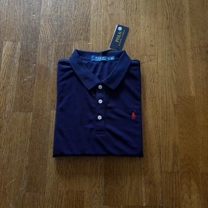 Marinblå Polo Ralph Lauren t-shirt - Snygg marinblå t-shirt från Polo Ralph Lauren med klassisk krage och tre vita knappar framtill. Ikonisk röd logga broderad på bröstet. T-shirten är kortärmad och gjord i mjuk bomull som känns skön mot huden. Perfekt för en chill och stilren look.