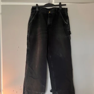 Svarta Carhartt cargopants wide fit - Grymma svarta cargopants från Carhartt med bred passform och slitna, fransiga benslut. Byxorna har flera stora fickor och klassisk Carhartt-logga bak. Perfekta för en avslappnad streetstil. Materialet är robust bomull.