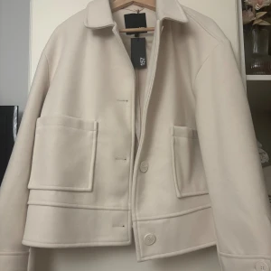 Beige ulljacka  - Stilren beige jacka från 157 i ullblandning. Jackan har klassisk krage, stora framfickor och knäppning med knappar framtill. Perfekt för dig som gillar minimalistisk och clean look. Passformen är normal och modellen är rak med långa ärmar.