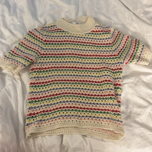 Randig stickad topp från Zara - Färgglad stickad topp från Zara med korta ärmar och hög krage. Tröjan har virkade ränder i vitt, rött, grönt, blått, gult och lila. Perfekt för dig som gillar retrovibbar och unika plagg. Säljes i storlek S.