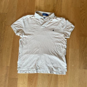 Beige slim fit piké från Polo Ralph Lauren - Snygg vit pikétröja från Polo Ralph Lauren i slim fit-modell. Klassisk krage med två knappar och den ikoniska broderade loggan på bröstet. Tillverkad i mjuk bomull som andas, perfekt för en clean och fräsch look.
