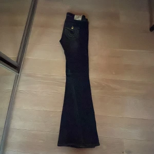 True Religion Joey Low Rise Flare Jeans - Mörkblå jeans från True Religion, modell Joey Low Rise Flare. Klassisk femficksdesign med kontrastsömmar och ikonisk fickdetalj bak. Bootcut/flare passform och låg midja. Tillverkade i slitstarkt bomullsjeansmaterial.