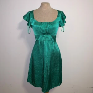 Grön klänning från Dunnes - Snygg grön kortklänning från Dunnes i glansigt material. Markerad midja med knytband bak och en djup rundad urringning fram. Perfekt för dig som gillar att sticka ut med färg och glans. Tyvärr är det lite trådar lösa (se sista bilden) men inget som syns på långt håll. Storlek 36.