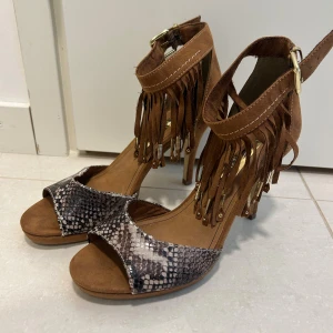 Bruna skorna med fransar och ormmönster - Snygga skorna med hög klack i brunt mocka och coolt ormmönster framtill. Skorna har öppen tå, ankelrem med spänne och fransdetaljer med guldfärgade metalldetaljer. Perfekta för dig som vill sticka ut med en boho-vibe.