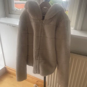 Vit/beige teddyjacka med huva - Supermjuk och fluffig vit teddyjacka med huva. Jackan har en enkel stängning framtill och är perfekt för kyliga dagar. Den har en oversized känsla och är helfärgad utan mönster. Passar dig som vill ha en mysig och trendig look.