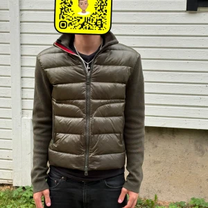 Olivgrön pufferjacka från Moncler - Säljer en olivgrön pufferjacka från Moncler med stickade ärmar och quiltad front. Jackan har hög krage, dragkedja och Moncler-logga på ärmen. Perfekt för höst och vinter, riktigt snygg och stilren modell som funkar till det mesta.