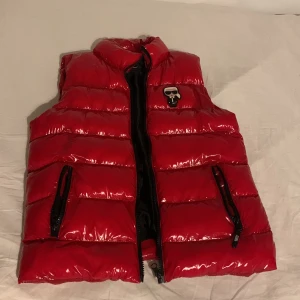 Röd dunväst från Karl Lagerfeld - Snygg röd dunväst från Karl Lagerfeld med glansig finish och svart foder. Västen har två fickor med dragkedja, hög krage och tryck med Karl Lagerfeld-figur på bröstet samt stor KARL-logga på ryggen. Perfekt för att sticka ut i mängden.