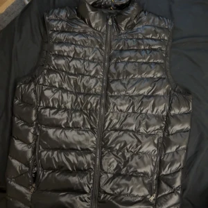 Svart dunväst från Polo Ralph Lauren - Snygg svart dunväst från Polo Ralph Lauren med quiltad design och hög krage. Västen har två fickor med dragkedja och en tydlig Polo-dragkedja framtill. Perfekt att slänga över en hoodie för en clean look. Hör gärna av dig vid funderingar!🤗