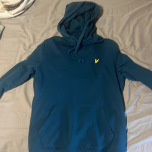 Blå hoodie från Lyle & Scott - Snygg marinblå hoodie från Lyle & Scott med klassisk gul logga på bröstet. Tröjan har huva med snörning och en stor magficka framtill. Perfekt för dig som gillar en clean och stilren look. Materialet är mjukt och skönt, passar till chill eller streetwear.