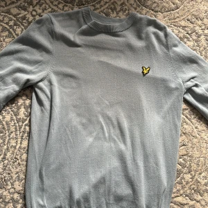 Ljusgrå stickad tröja från Lyle & Scott - Säljer en stilren ljusgrå stickad tröja från Lyle & Scott med deras klassiska gula fågel-logga på bröstet. Tröjan har rund halsringning och långa ärmar, perfekt för lager-på-lager. Skön och enkel att matcha till olika outfits. Använd 1 gång 