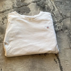 Vit t-shirt från Gant med logga - Vit t-shirt från Gant i klassisk modell med rund hals och korta ärmar. Liten broderad Gant-logga med sköld på bröstet. Tillverkad i mjuk bomull som känns skön mot huden. Perfekt för en clean och enkel stil.