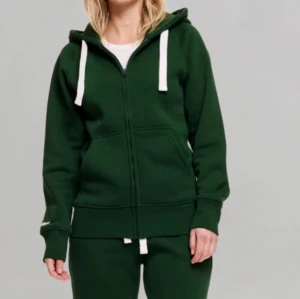 Russemerch zip up Hoodie - Säljer en mörkgrön hoodie från russemerch med vit snörning i huvan och dragkedja framtill. Tröjan har långa ärmar, ribbade muddar och fickor på sidorna. Perfekt för chill dagar eller när du vill vara extra bekväm. Den har någon liten fläck fram på min inget man tänker på.