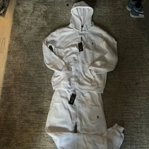 Vit polo tracksuit M - Vit hoodie från Polo Ralph Lauren i storlek M. Klassisk design med dragkedja, huva och två fickor framtill. Liten broderad logga på bröstet. Tillverkad i mjuk bomull, perfekt för en clean och avslappnad stil.