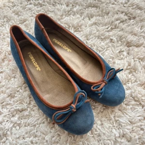 Blå mocka ballerinaskor från Penelope - Supersöta ballerinaskor i blå mocka från Penelope med bruna detaljer och rosett framtill. Skorna har rund tå och platt sula, perfekta för en chill och stilren look. Bruna kanter ger en snygg kontrast till den blå färgen. Köpt begagnat men är i super bra skick! Säljer då de för stora 