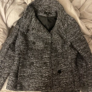 Svartvit rutig skepparkavaj från H&M - Säljer en snygg skepparkavaj från H&M i svartvitt rutigt mönster. Jackan har dubbelknäppning med svarta knappar, bred krage och är i en lite kortare modell. Materialet känns tjockt och strukturerat, perfekt för dig som gillar klassisk stil med en modern twist.