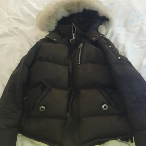 Svart Moose Knuckles dunjacka med päls - Säljer en svart dunjacka från Moose Knuckles med fluffig päls runt huvan och ikonisk logga på ärmen. Jackan har flera fickor med dragkedja och snygga metalldetaljer. Perfekt för kalla vinterdagar och riktigt varm.