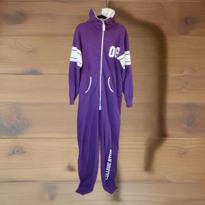 Lila onesie hoodie från College Style - Lila onesie med vit dragkedja och huva från College Style. Snygga vita detaljer på ärmarna och siffran 06 på bröstet. Mjukt bomullsmaterial, två fickor framtill och ribbade muddar vid ärmarna. Har blivit lite rosaspräcklig på baksidan.