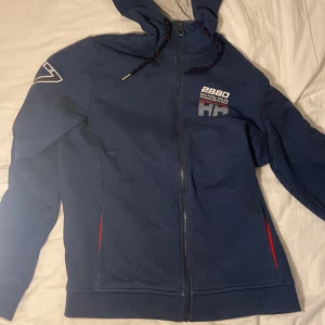 Marinblå Helly Hansen hoodie med dragkedja - Marinblå hoodie från Helly Hansen med hel dragkedja, broderad text och HH-logga på bröstet samt siffran 77 på ärmen. Tröjan har huva med snörning, röda detaljer vid fickorna och är fodrad i grått tyg på insidan av huvan. Perfekt för chill dagar.
