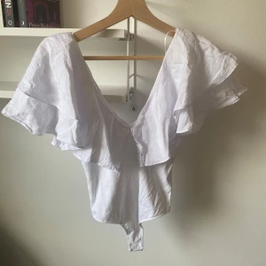 Vit volangbody från Zara - Supersnygg vit body från Zara med stora volanger över axlarna och djup v-ringning. Modellen är ärmlös och har en feminin, omlottliknande känsla. Perfekt att styla med jeans eller kjol för en trendig look🌷