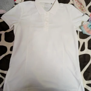 Vit piké t-shirt från Craft - Vit piké t-shirt från Craft i storlek S. Klassisk modell med krage, tre knappar framtill och diskret logga på ärmen. Tillverkad i ett lätt och funktionellt material som andas, perfekt för sportiga dagar eller en clean stil.