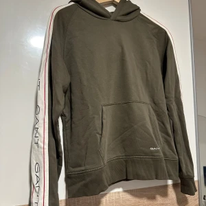 Olivgrön hoodie från GANT med logga - Snygg olivgrön hoodie från GANT med vita ärmar och röda detaljer längs sidorna. GANT-logga på bröstet och längs ärmarna. Klassisk känguruficka framtill och huva. Perfekt för en avslappnad och sportig stil. Endast använd engång.