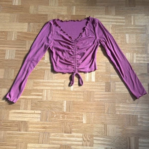 Röd croppad ribbad topp med snörning - Säljer en vinröd (bilden ger en mer lila ton men i verklighet mer röd) croppad topp med långa ärmar och v-ringning. Toppen har ribbat tyg, volangkant vid hals och ärmslut samt snörning framtill som ger en snygg rynkad effekt.