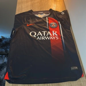 PSG matchtröja Nike blå/röd - Paris Saint-Germain fotbollströja från Nike med Qatar Airways-tryck. Tröjan är marinblå med röd och vit vertikal rand, PSG-emblem på bröstet och Dri-FIT-material som andas. Klassisk passform med korta ärmar och rund hals.