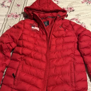 Röd pufferjacka med huva - Riktigt snygg röd pufferjacka från Polo Club med broderad logga på bröstet och huva. Jackan har dragkedja framtill och två sidofickor med dragkedja. Perfekt för kyliga dagar och ger en sportig vibe.