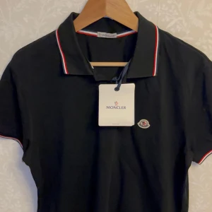 Svart  pikétröja med logga - helt ny och oandvänd Svart pikétröja  med taggen på och klassisk krage och knäppning framtill. Röd och vit rand längs krage och ärmslut, samt broderad logga på bröstet. Tillverkad i bomull för en skön och stilren look. INTE RIKTIG