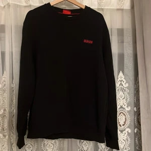 Svart sweatshirt från HUGO - Svart sweatshirt från HUGO med röd logga på bröstet. Klassisk rund halsringning och långa ärmar. Tröjan har en clean look och är tillverkad i mjuk bomull för maximal komfort. Perfekt för dig som gillar stilrena och snygga plagg.