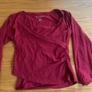 Vinröd omlottopp från Bondelid - Säljer en vinröd omlottopp från Bondelid i storlek S. Toppen har långa ärmar, v-ringning och en snygg omlottdesign som ger en schysst siluett. Tillverkad i mjuk bomull som känns skön mot huden. Perfekt att styla med jeans eller kjol.