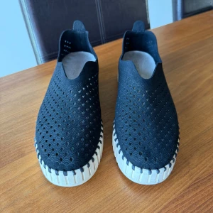 Ilse Jacobsen svarta slip-on skor - Svarta slip-on skor från Ilse Jacobsen med perforerat mönster och vit, räfflad sula. Skorna har rund tå och är tillverkade i syntetmaterial med snygga detaljer på hälen. Perfekta för dig som gillar stilren och modern design. 