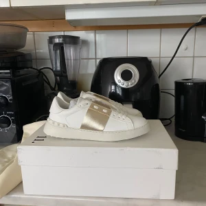 Valentino Garavani sneakers vit/guld - Snygga sneakers från Valentino Garavani i vitt skinn med bred metallic guldrem över vristen. Klassisk rund tå, platt sula och vita snören. Ikoniska nitar baktill på sulan ger extra edge. Perfekta för dig som vill sticka ut med lyxig streetstyle.