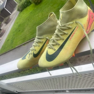Nike Mercurial fotbollsskor gul - Säljer ett par Nike Mercurial fotbollsskor i ljusgul färg med svarta swoosh-loggor och orange detaljer på hälen. Skorna har snörning, hög krage och platt sula med dobbar. Perfekta för dig som vill sticka ut på planen. Snygg och modern design!