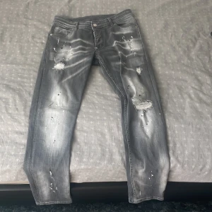 Grå Dsquared2 jeans med slitningar - Säljer ett par grå jeans från Dsquared2 med coola slitningar, hål och färgstänk för en edgy look. Jeansen har fem fickor, raka ben och en stor vit Dsquared2-patch bak. Perfekta för dig som gillar streetstyle och vill sticka ut.