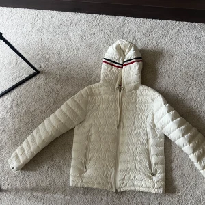 Vit Moncler dunjacka med huva - Säljer en snygg vit dunjacka från Moncler med huva och dragkedja framtill. Jackan har två fickor med dragkedja och en broderad Moncler-logga på ärmen. Huvan har en röd och marinblå rand som detalj. Perfekt för kyliga dagar!