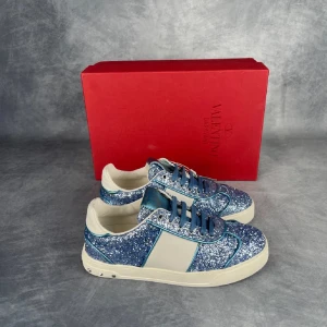 Valentino Flycrews glittriga sneakers - Säljer ett par blå glittriga Valentino Garavani sneakers med vita detaljer och coola nitar på hälen. Skorna är i storlek 37. Skorna har snörning, rund tå och en vit sula med camomönster undertill. Perfekta för dig som vill sticka ut med stil! Levereras med originalkartong.