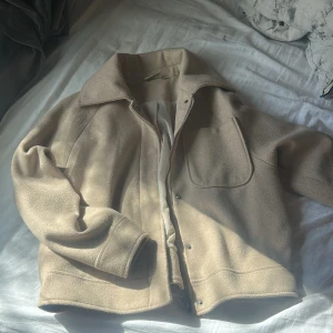 Beige tjockare kappa från Ullared , stl 36/38 - Snygg beige jacka från Ullared i ullblandning, storlek 36/38. Jackan har en clean look med stor krage och dragkedja och tryckknappar framtill samt en stor ficka på bröstet. Perfekt för dig som gillar minimalistisk och stilren design.