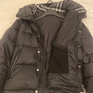 Svart pufferjacka från Burberry - Svart pufferjacka från Burberry med klassiskt rutigt foder i huvan och tydlig logga på bröstet. Jackan har dragkedja, tryckknappar och flera fickor. Perfekt för kalla dagar med sin vadderade design och snygga detaljer. Den har dock ett litet hål som går och fixa via en skräddare 