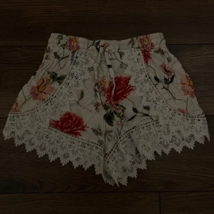Blommiga shorts med spetsdetaljer - Supersöta vita shorts med blommigt mönster i rött, rosa och grönt. Shortsens kanter är dekorerade med bred, vit spets och de har snörning i midjan. Perfekta för sommarens varma dagar och ger en romantisk vibe.