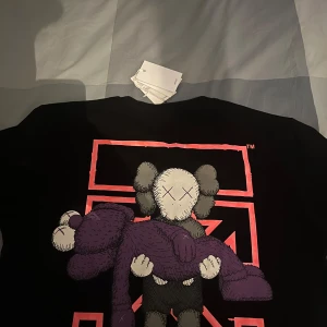 Svart Off-White x Kaws t-shirt XL - Svart t-shirt från Off-White i samarbete med Kaws, storlek XL. Framsidan har ett litet tryck med texten 'Off-White Kaws' och baksidan har en stor grafisk illustration med Kaws-figurer i lila och grått. Klassisk rund hals och korta ärmar. Materialet är mjuk bomull.