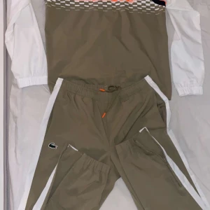 Lacoste tracksuit - Säljer ett par beige träningsbyxor från Lacoste med vita sidor och dragsko i midjan. Byxorna har en liten Lacoste-logga på vänster ben och orange detaljer vid snörningen. Perfekta för sport eller chill.